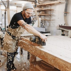 Carpenter working with electric planer on wooden plank in workshop.